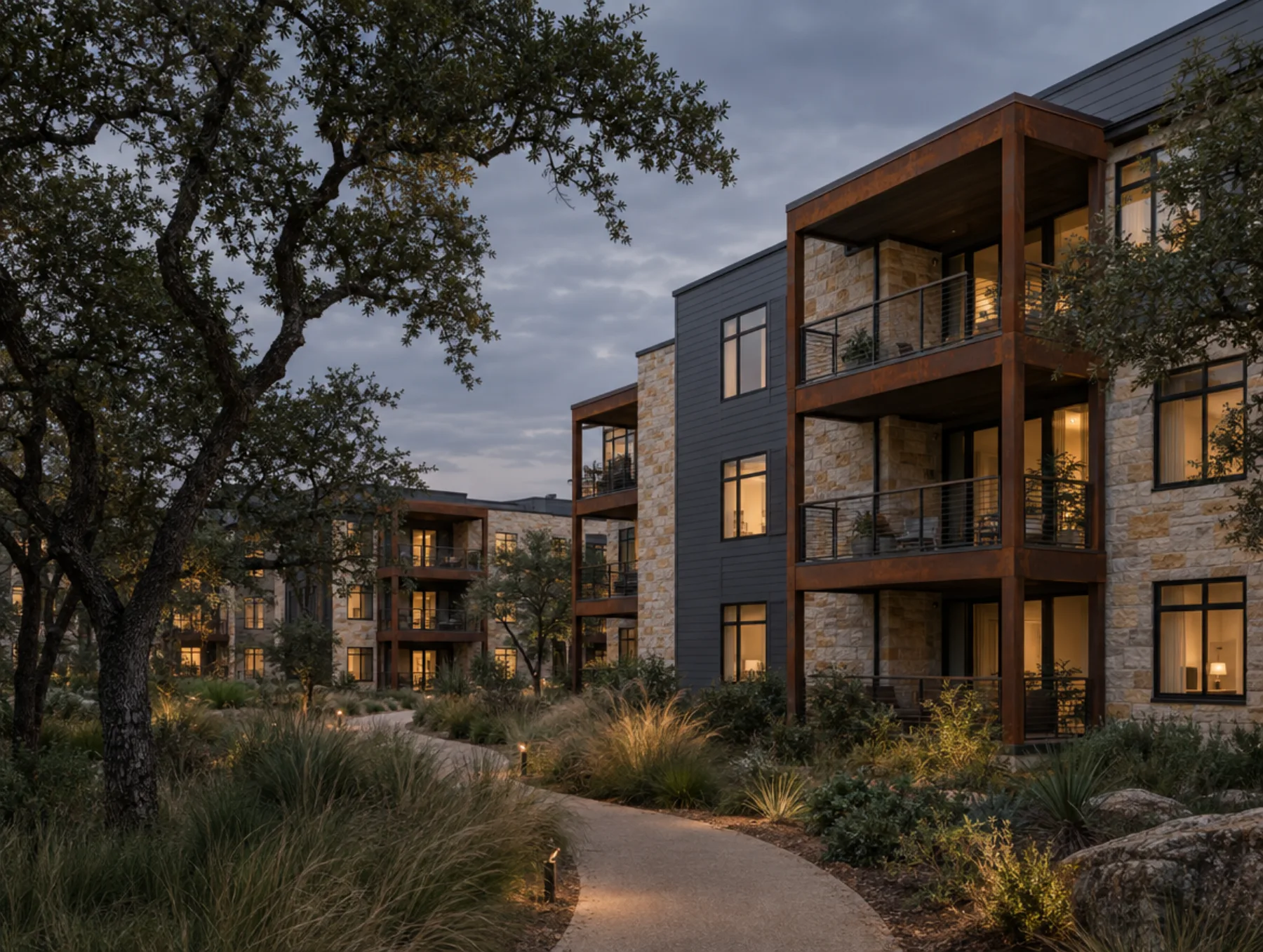 A stone and weathered-steel apartment community along a winding path under live oaks at dusk